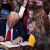 Donald Trump in a suit at a desk signing an executive order, surrounded by women and girls