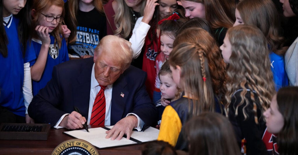 Donald Trump in a suit at a desk signing an executive order, surrounded by women and girls