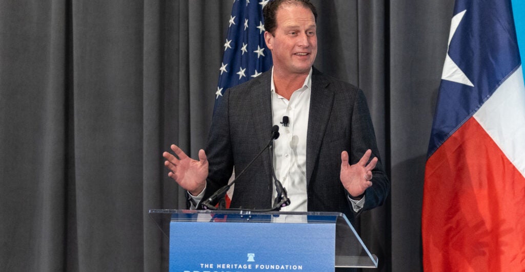 Texas Rep. August Pfluger wears a black suit and white tee shirt while standing at a pulpit.