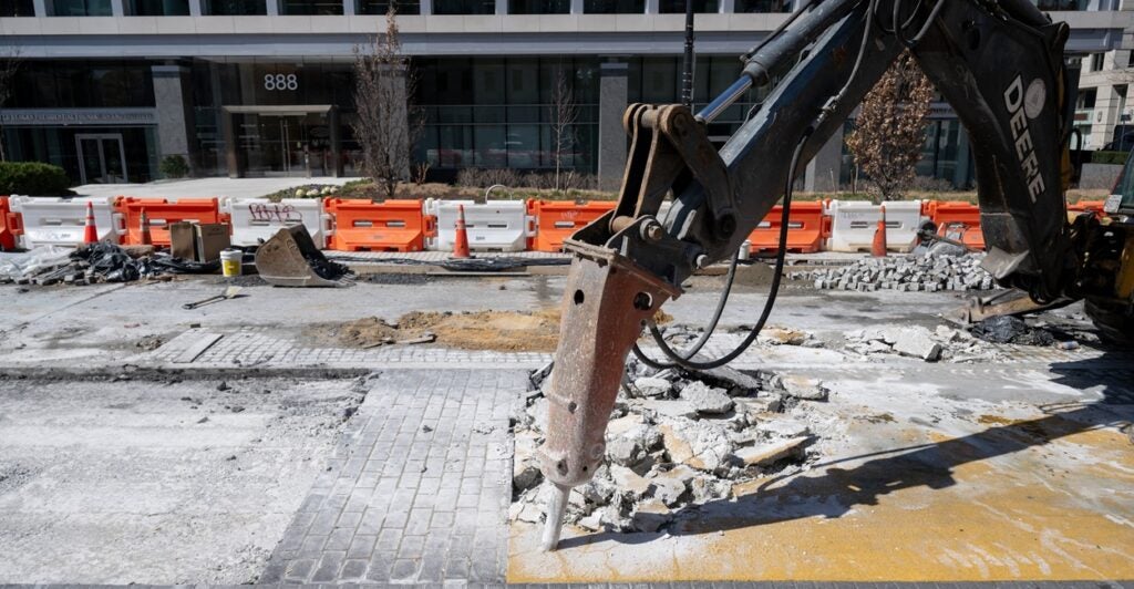 An excavator jackhammer digs into pavement as broken pieces of concrete lay on the ground.