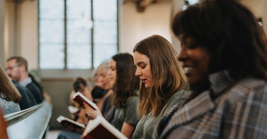 Women are sitting in a pew while reading a hymn book in a religious facility.