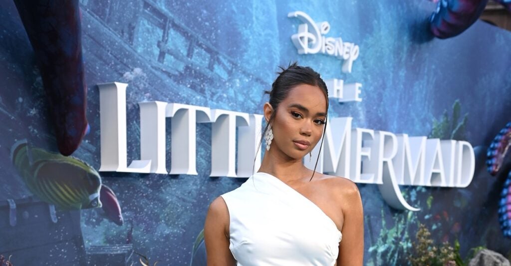 Michelle Domingos is wearing a white dress with her hands at her side as she stands in front of a sign for Disney's "Little Mermaid."