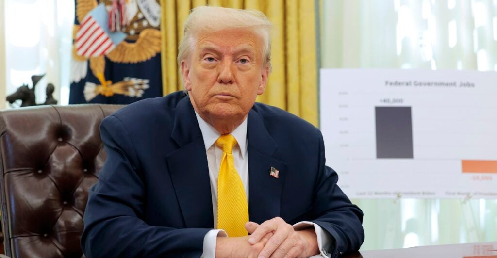 Donald Trump sits at a desk with his hands folded in front of him as he looks straight forward.