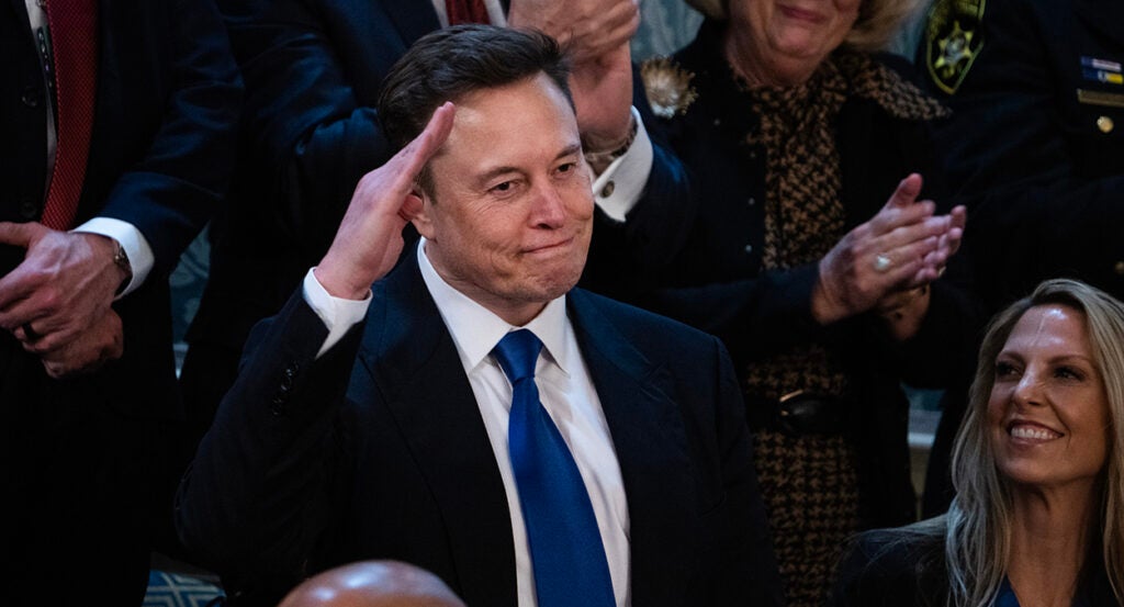 Elon Musk salutes while wearing a suit with a blue tie.