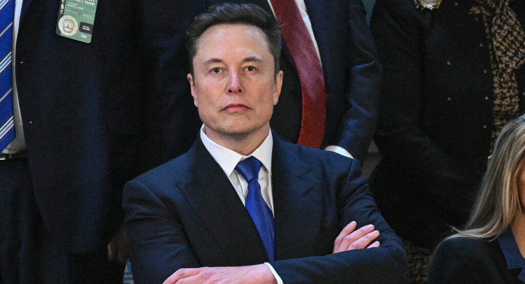 Elon Musk in a suit with a blue tie stands resolute