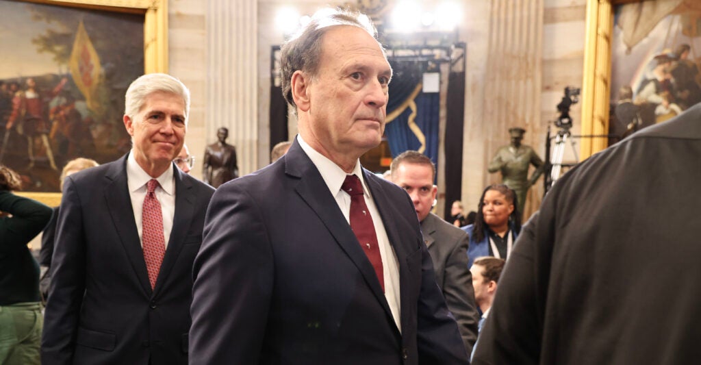 Samuel Alito stands wearing a slate grey suit and a dark red necktie.