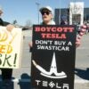 Two people stand side by side holding up signs protesting Elon Musk and Tesla.