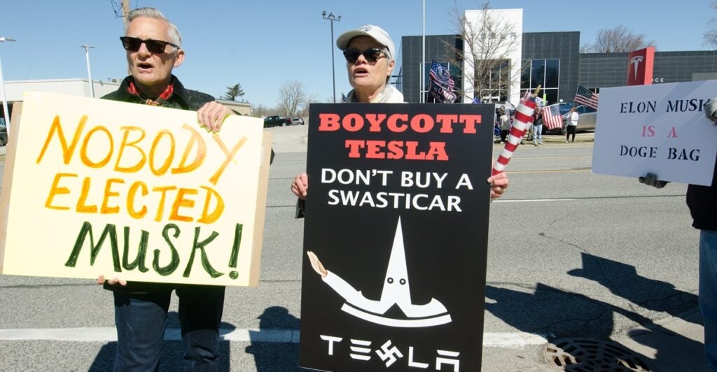 Two people stand side by side holding up signs protesting Elon Musk and Tesla.