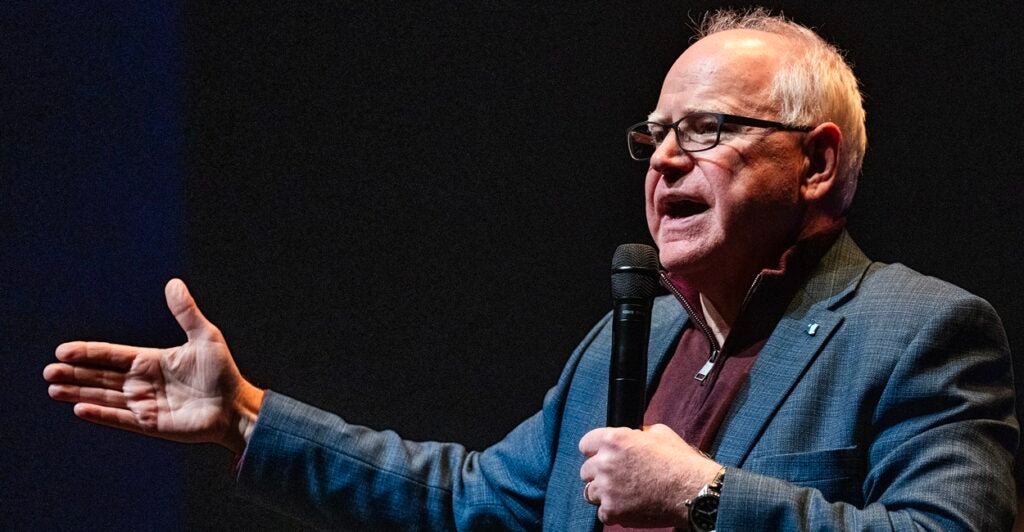 Tim Walz stands with a microphone in his left hand and his right hand reaching out as he speaks to an audience.