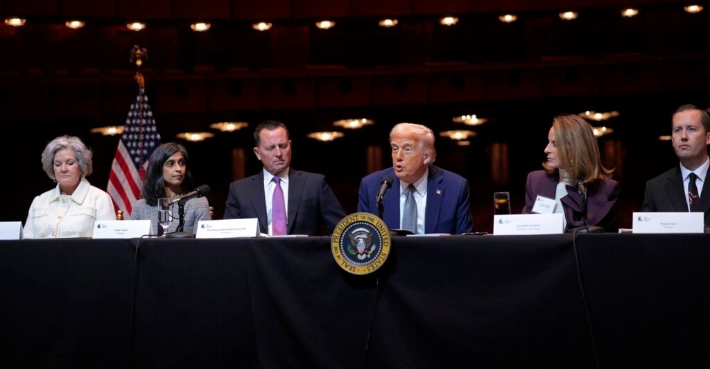 Donald Trump sits at a table and looks out at reporters with both arms at his side.