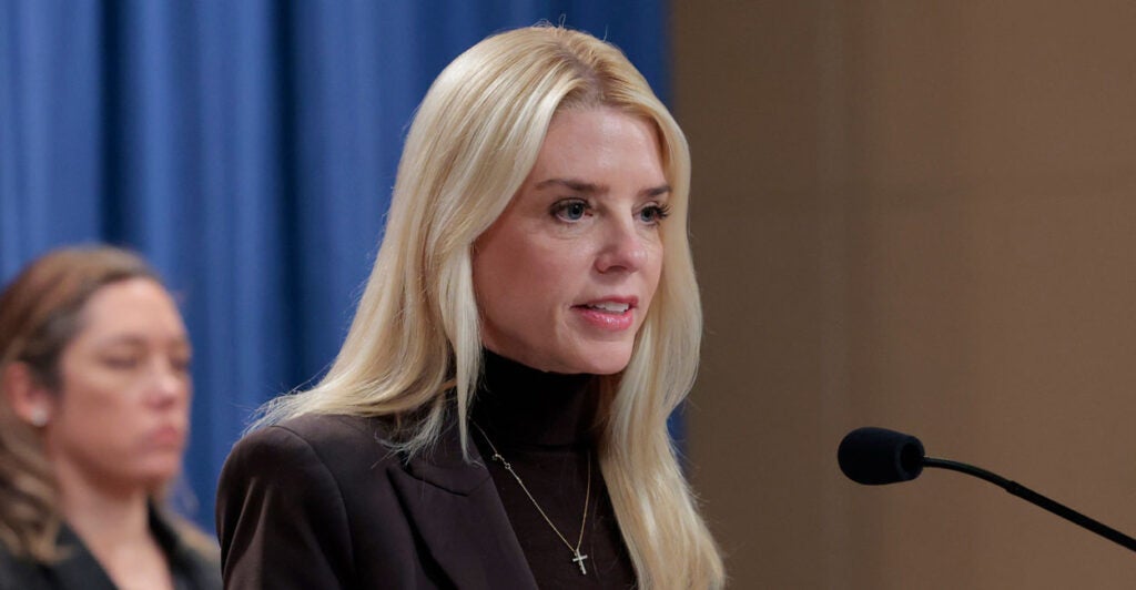 Attorney General Pam Bondi in a brown turtleneck and brown jacket speaking at a podium