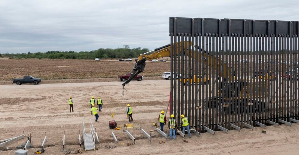 A large excavator begins to dig into the ground near where a wall is being built along the southern border.