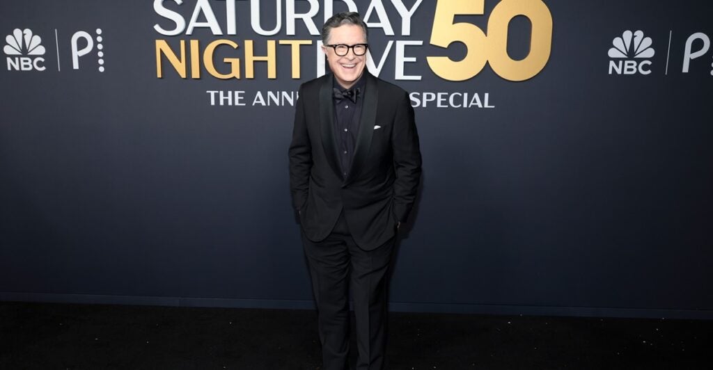 Stephen Colbert stands with his hands in his pockets oni the red carpet.