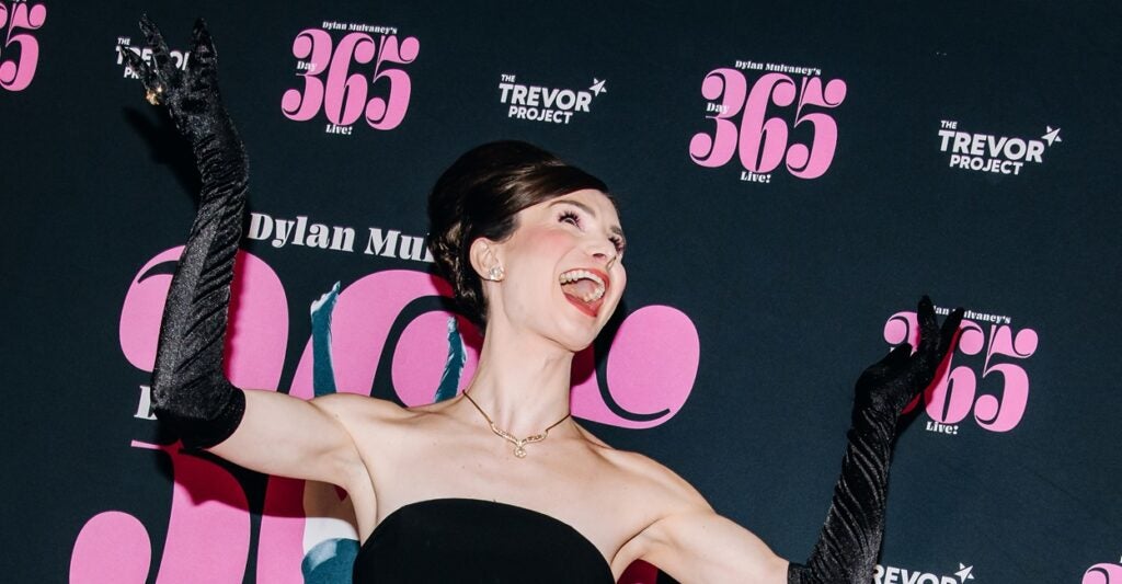 Dylan Mulvaney has both hands raised and is reacting with mouth open while on the red carpet.