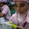 An older Chinese man in a Chinese e-cigarette manufacturer tests an e-cigarette with vapor streaming out of his nose.