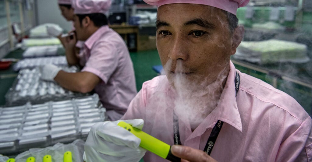 An older Chinese man in a Chinese e-cigarette manufacturer tests an e-cigarette with vapor streaming out of his nose.
