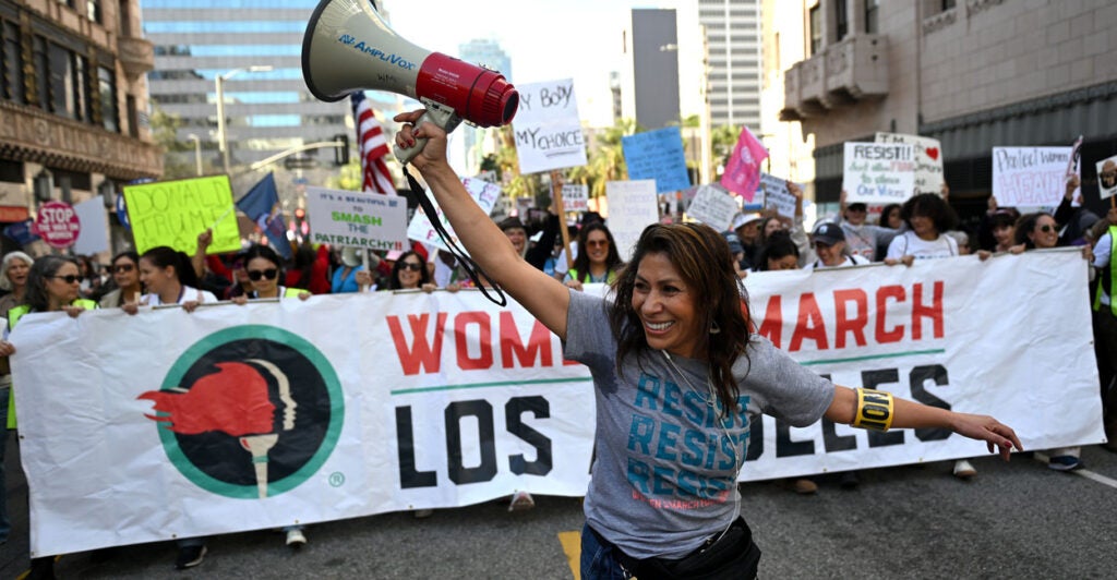 a woman raising her hands with a megaphone leads a protest march through the streets of Los Angeles with women with protest signs and holding a large "Women's March Los Angeles" banner.
