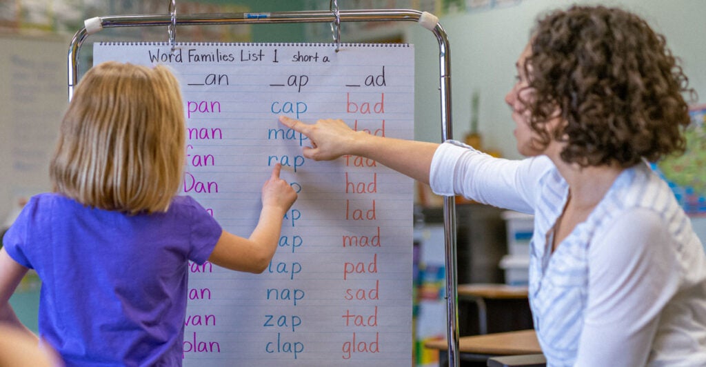 a six-year-old girl in a purple top and her teacher in a white top point to a chart with simple words on it like cat and hat