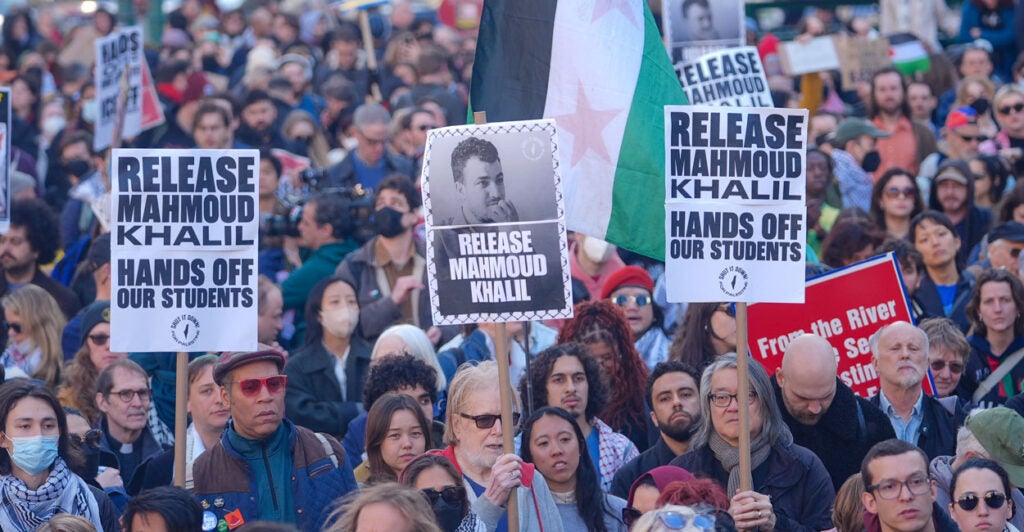 crowds of protesters, young and old, hold up a Palestinian flag and placards saying Release Mahmoud Khalil
