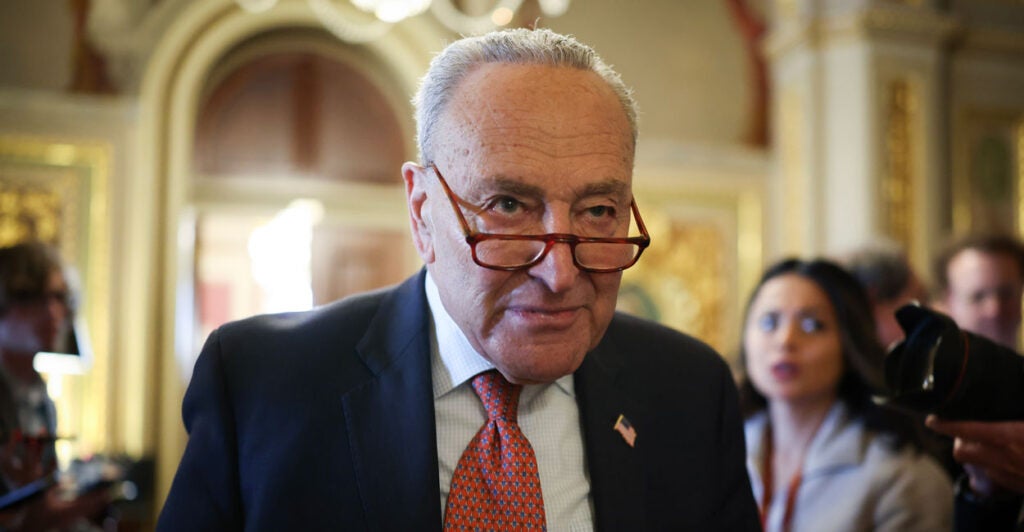 Chuck Schumer in a dark suit and red tie with glasses down his nose looks intently ahead