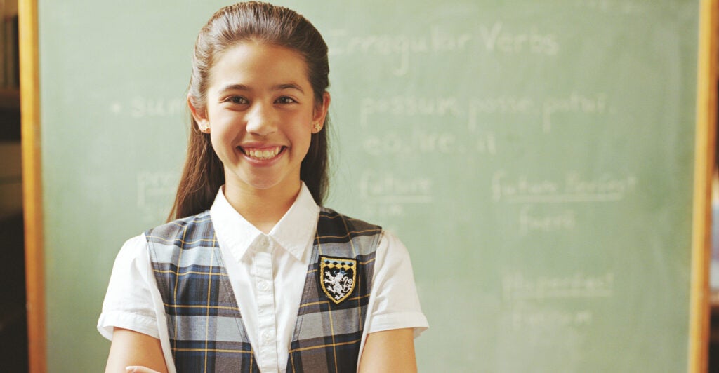 A young girl, wearing private-school attire, stands with arms crossed in front of a chalkboard.