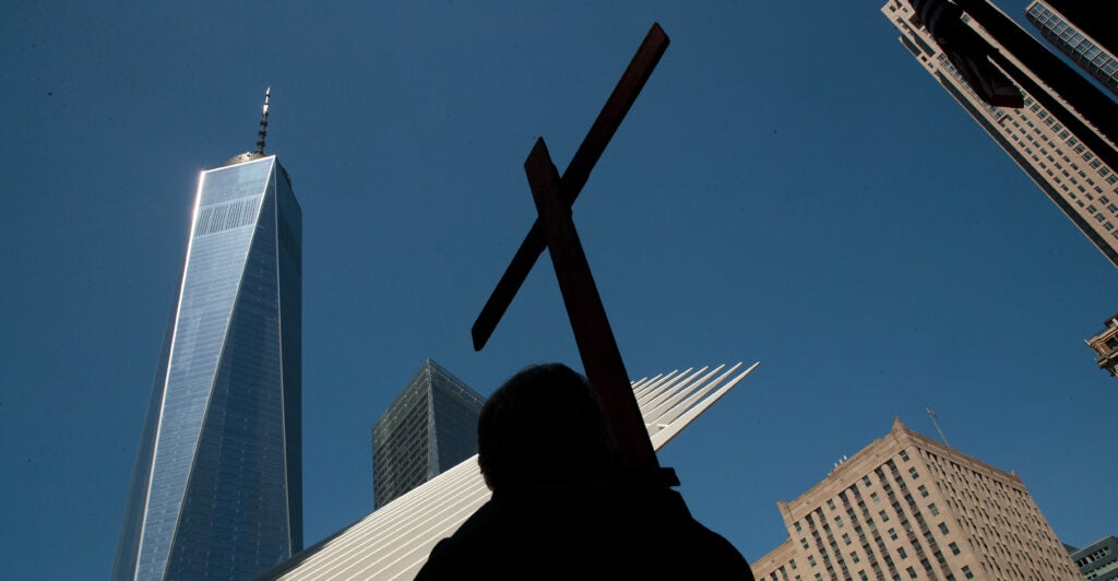 A low-angle shot of a man in silhouette carrying a cross, against a backdrop of the Freedom Tower other high-rises and a perfect blue sky.