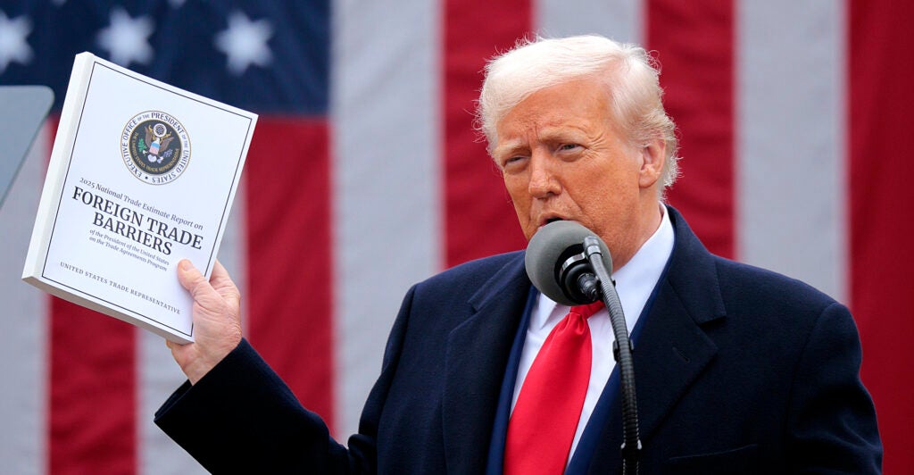 President Trump stands before a large American flag while speaking and holding up a copy of the 2025 National Trade Report.