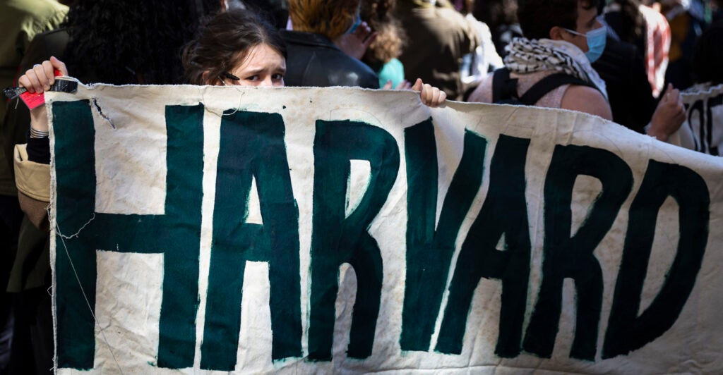 Mostly obscured students hold a green lettered "Harvard" sign.