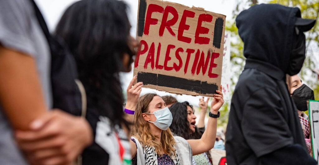 Masked protesters at a Pro-Palestinian rally. A white woman holds up a hand-drawn in blood red sign reading "Free Palestine."