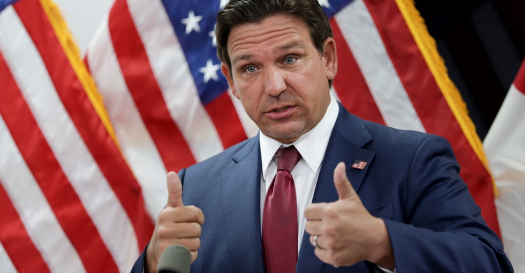 Florida Governor Ron DeSantis and a blue suit and red tie with American flags in the background gives two thumbs up