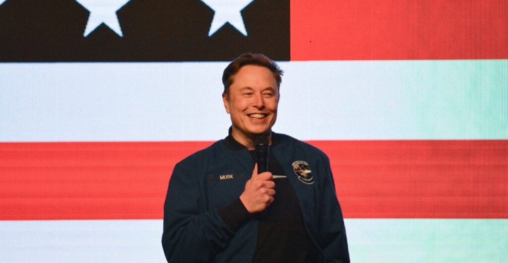 Elon Musk stands with a microphone in his hand and smiles with the backdrop of an American flag.