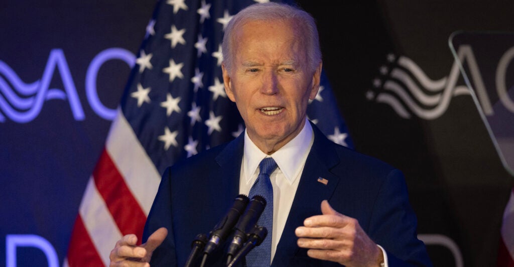 former President Joe Biden in a blue suit and blue tie speaking at a podium with an American flag in the background