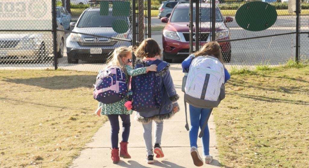 Three little schoolgirls wearing backpacks, about eight-years-old walk toward a gate to leave school. Blonde girl on left has arm draped on back of the middle girl.