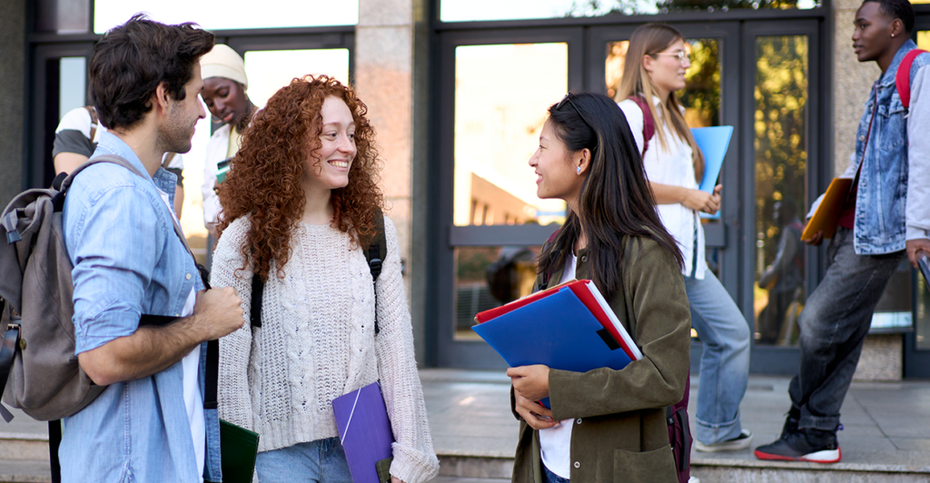 college students stand outside of the school building with notebooks and backpacks talking with one another