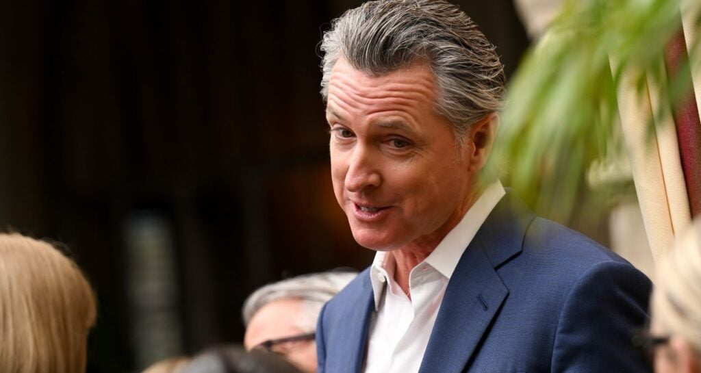 California Gov. Gavin Newsom, in a blue jacket and open-collar white shirt, speaks, eyebrows raised.