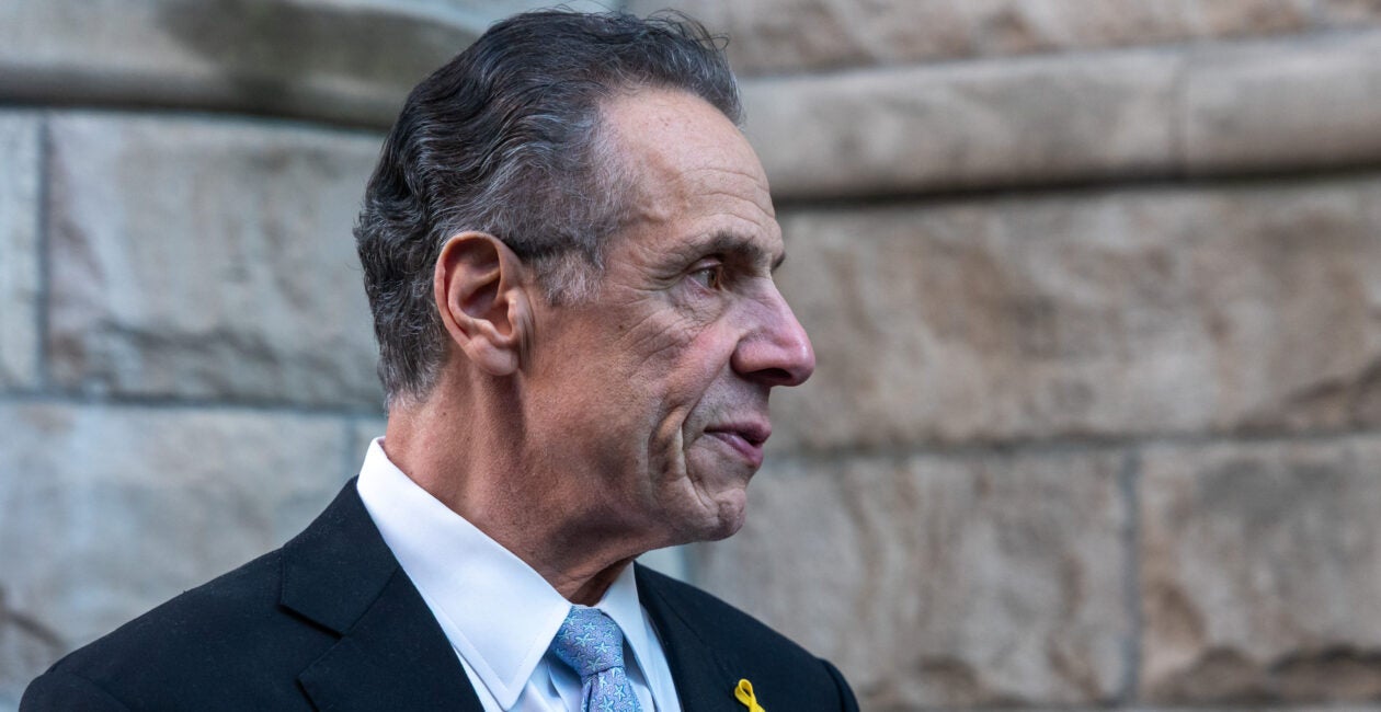 Andrew Cuomo stands wearing a black suit and light-blue necktie.