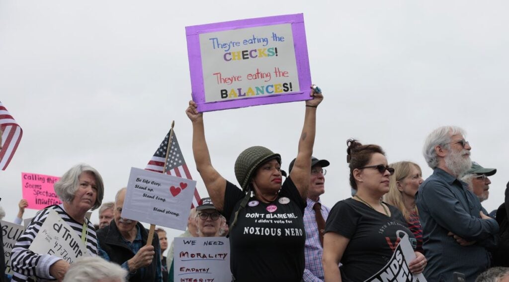 Black woman holds up a sign reading "They're eating the checks, they're eating the balances" amid crowd of other protesters anti-Trump "Hands Off" rally in Washington, DC."