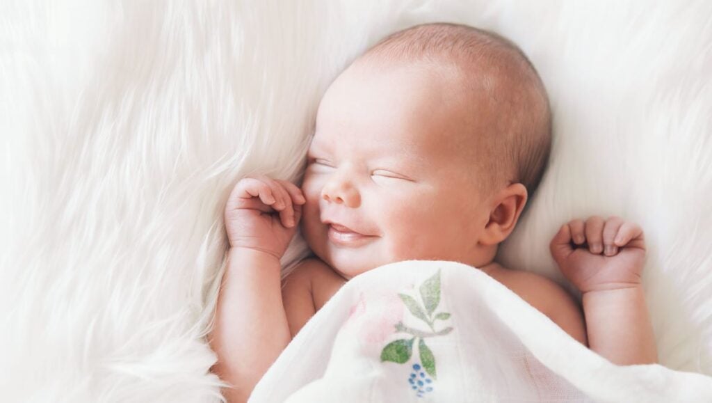 Adorable smiling newborn asleep on white blanket, while covered in a blanket with leaves and a flower.