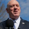 A smiling Tom Homan in a dark suit speaks into a microphone outdoors to reporters not seen in the photo
