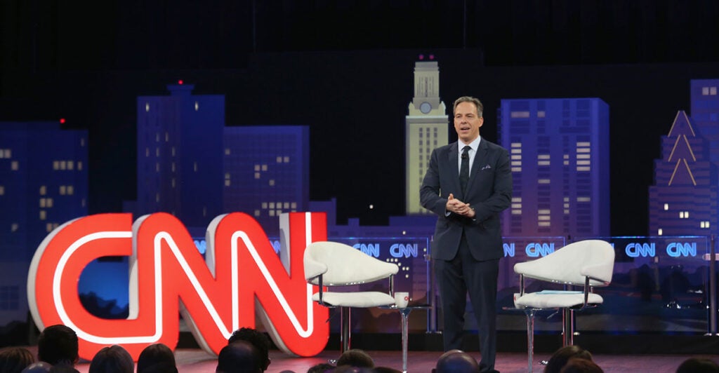 Jake Tapper standing in a black suit on a stage in front of a red CNN sign.