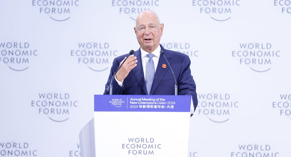 Klaus Schwab in a blue suit in front of a sign reading "World Economic Forum"