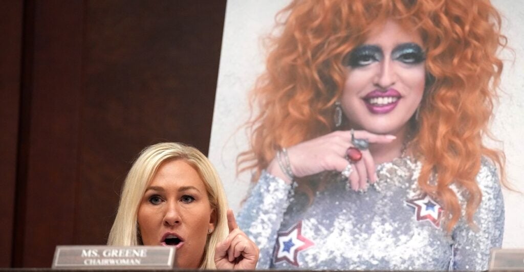 Just the head of Marjorie Taylor Greene can be seen above a desk while in the background is a large photo of a drag queen.