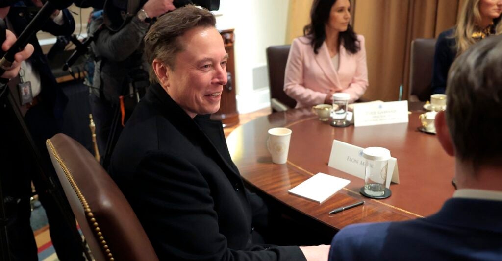 Elon Musk sits in a chair at a table and smiles as his head is turned to his right.