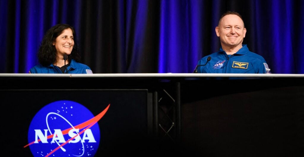 Butch Wilmore smiles as he sits at a table with fellow astronaut Suni Williams to his right.