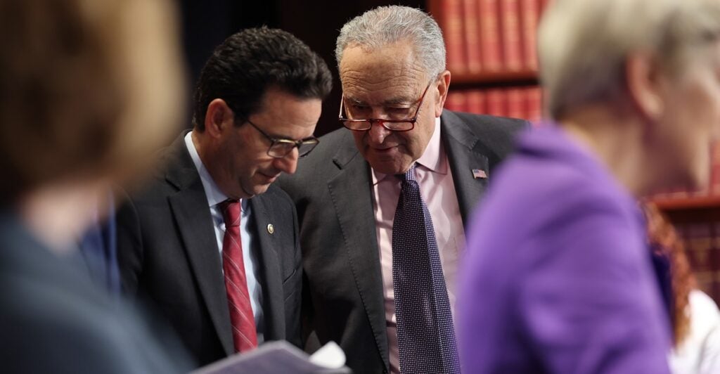 Brian Schatz puts his head down as he listens to Chuck Schumer while standing next to him.