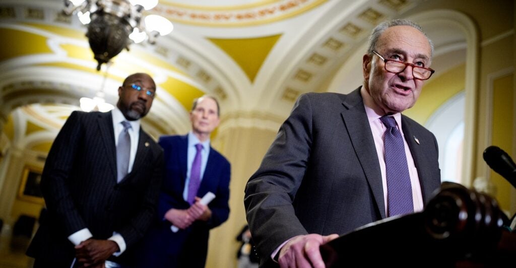 Chuck Schumer stands at a podium with his right hand gripping the podium as he prepares to speak to reporters.