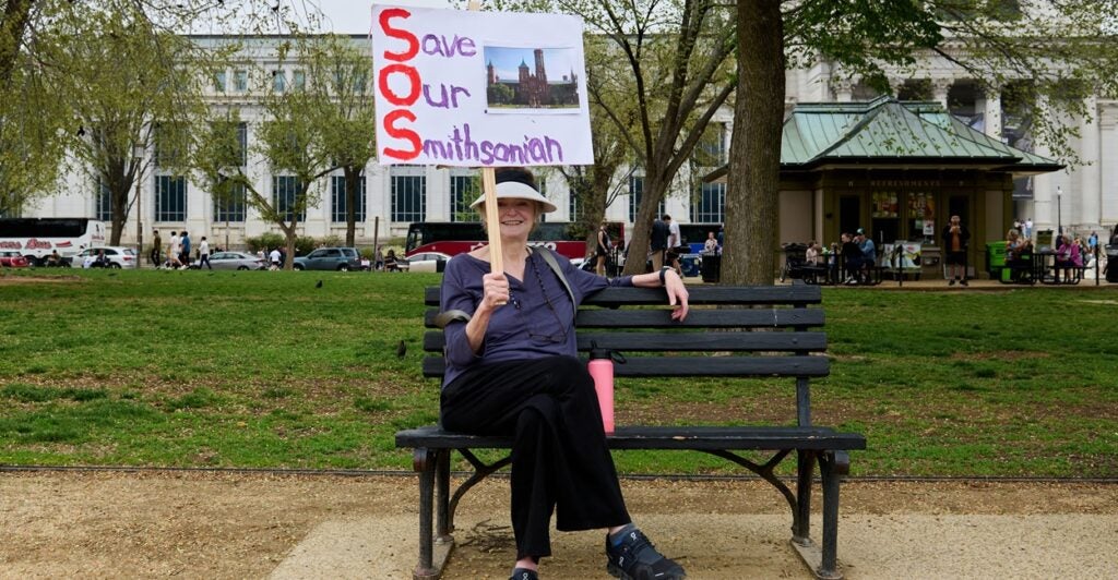 A woman sits on a park bench and holds a sign up in the air with her right hand. The sign states, "Save Our Smithsonian".