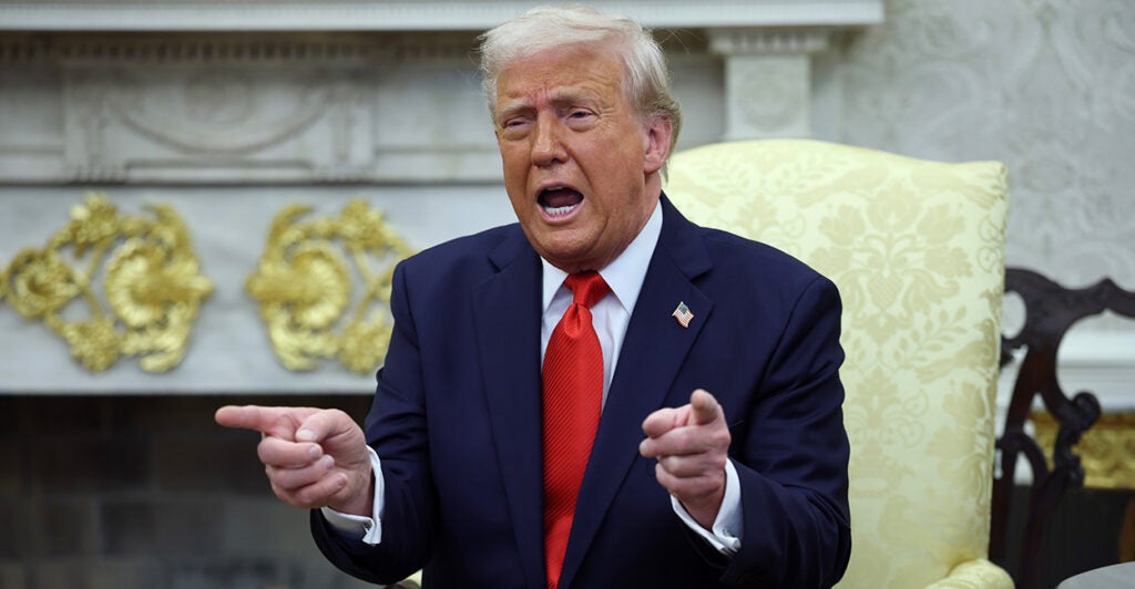 President Donald Trump sits in the Oval Office in a yellow chair wearing a dark blue suite and red tie. He is speaking and both his finders are pointed forward.