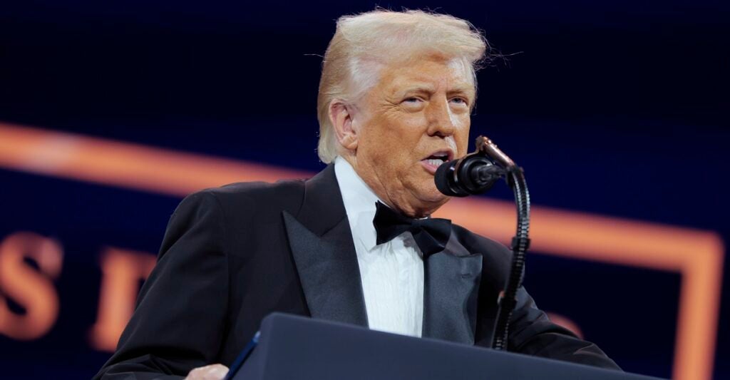 Donald Trump wears a tuxedo as he stands in front of a podium and speaks into a microphone.