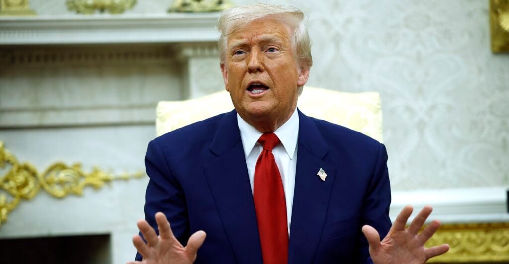 Donald Trump sits in a chair and raises both his hands while talking.